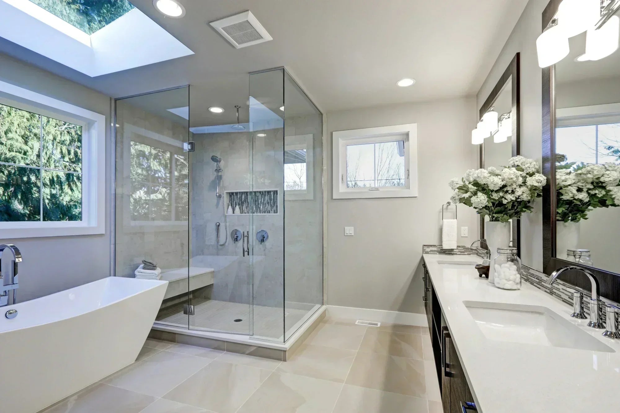 Modern bathroom with large-format stone-effect floor tiles, walk-in glass shower, freestanding bath and double vanity with mirrors and skylight.