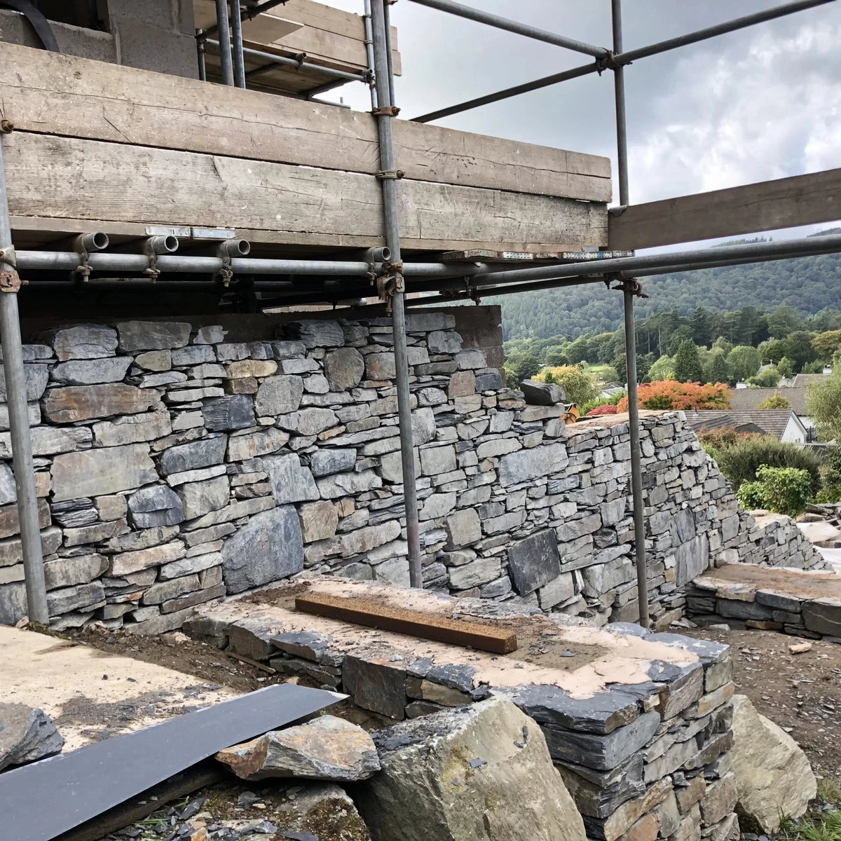Stone wall under construction with scaffolding and wooden planks, surrounded by greenery and mountains.