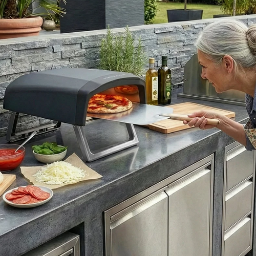 Person preparing pizza on an outdoor pizza oven in a backyard setting.