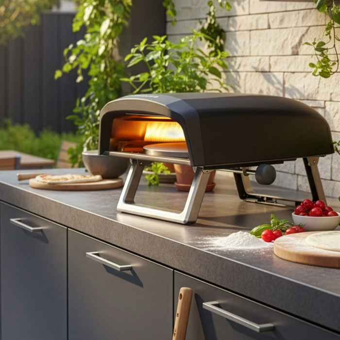 Outdoor kitchen scene with a pizza oven on a countertop, surrounded by garden furniture and plants.